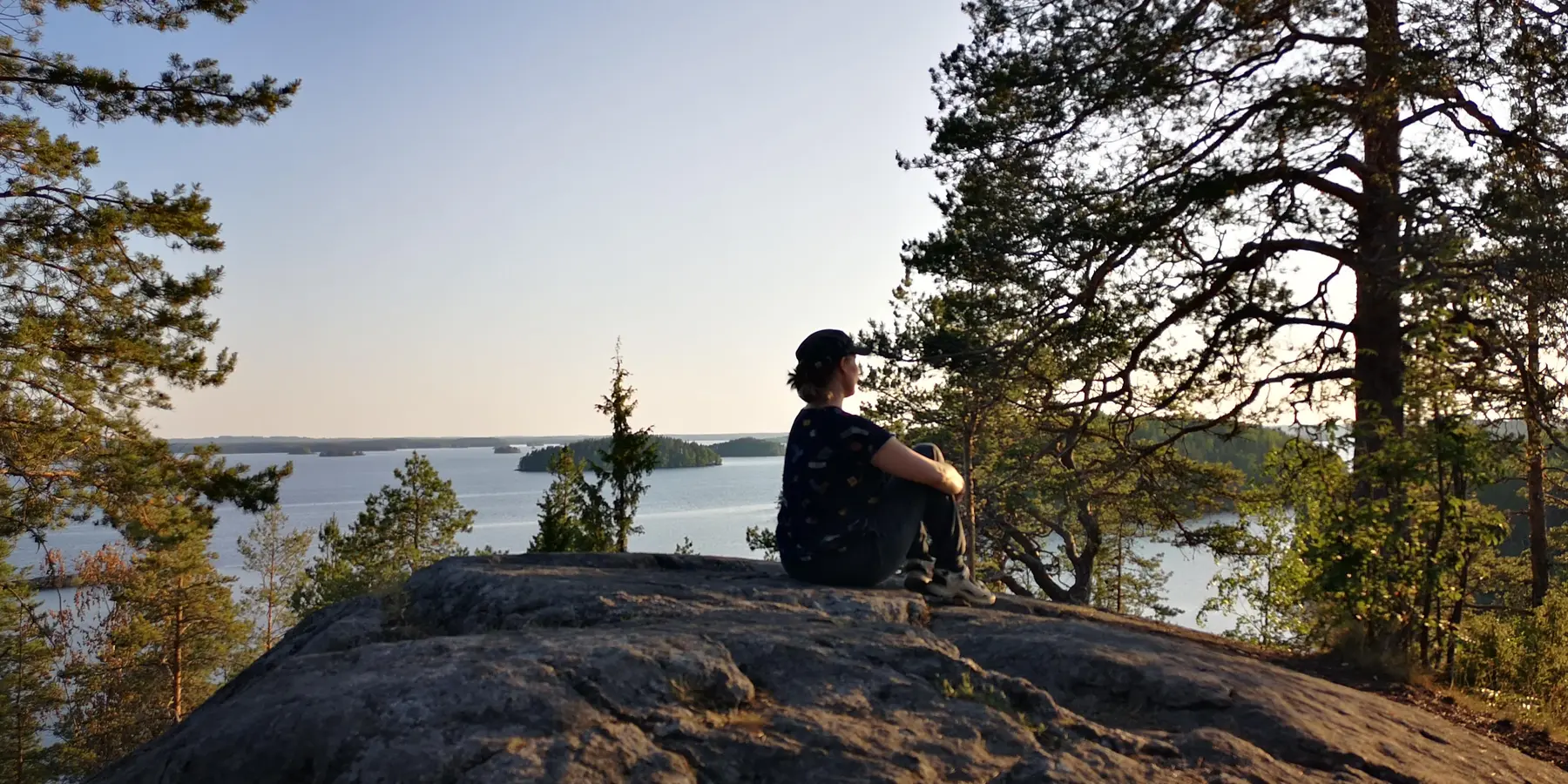Petra istuu kalliolla ja katselee järvimaisemaa